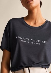 Dunkelgraues T-Shirt mit kurzen Ärmeln und rundem Ausschnitt, versehen mit dem weißen Drucktext: "RUE DES SOURIRE - PARIS, FRANKREICH." Weicher Stoff.