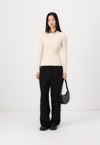 Cream ribbed sweater with long sleeves, paired with black wide-leg trousers. Model holds a black, perforated handbag. Smooth texture, fitted design.