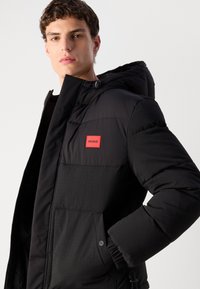 Giubbotto piumino nero con cappuccio, caratterizzato da una texture liscia, un patch logo HUGO rosso e polsini elastici. Include chiusure con zip e bottoni.