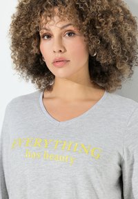 Graues V-Ausschnitt-T-Shirt mit gelbem gesticktem Text "ALLES hat Schönheit." Weicher, strukturierter Baumwollstoff, lockerer Schnitt und lange Ärmel.