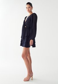 Mornarsko modra obleka z dolgimi naborkami, globokim V-izrezom, nabranim robom in izreznimi podrobnostmi, v kombinaciji s prozornimi sandali na peto.