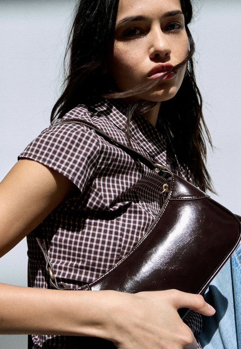 Jeune femme aux longs cheveux foncés portant une chemise à carreaux tient un sac à bandoulière en cuir marron brillant sur un fond clair.