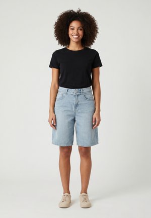 Femme aux cheveux bouclés portant un t-shirt noir, un short en jean bleu clair et des baskets beiges, debout et souriante sur un fond uni.