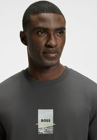 Sudadera gris de canalé con un gráfico central que presenta la palabra "BOSS" y un acento de impresión fotográfica. Cuello redondo y mangas cortas.