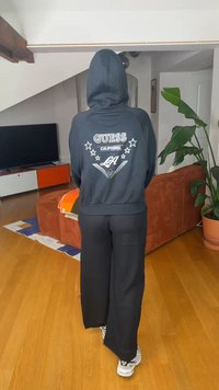 Personne portant un sweat à capuche noir Guess avec le texte "California LA" au dos, pantalon noir et baskets blanches, debout dans un salon.