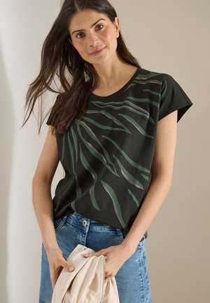 Femme aux longs cheveux foncés portant un t-shirt noir avec des motifs de feuilles vertes et un jean bleu, tenant une veste beige pliée.