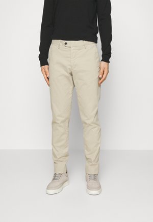 Osoba ubrana w beżowe spodnie o prostym kroju, jasnoszare trampki oraz czarną bluzkę z długim rękawem, stojąca na tle jednolitego, jasnego tła.