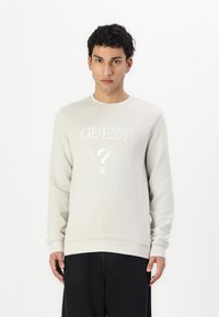 Sudadera gris clara de material suave, con cuello redondo y mangas largas. Logo bordado y elementos de diseño en la parte delantera.