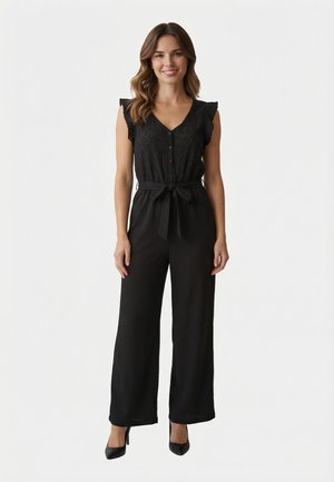 Donna in piedi con tuta nera senza maniche con bottoni, cintura annodata in vita e scarpe nere con tacchi alti, su sfondo semplice.