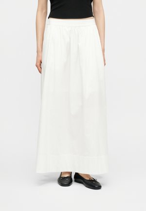 Eine Frau trägt einen langen weißen Rock mit gerafftem Bund, ein schwarzes bauchfreies Oberteil und schwarze Ballettschuhe und steht vor einem einfarbigen Hintergrund.