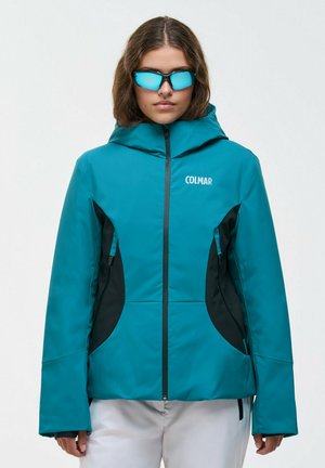 Une femme porte une veste de ski Colmar bleu sarcelle avec des panneaux latéraux noirs et des lunettes de soleil bleues réfléchissantes sur un fond uni.