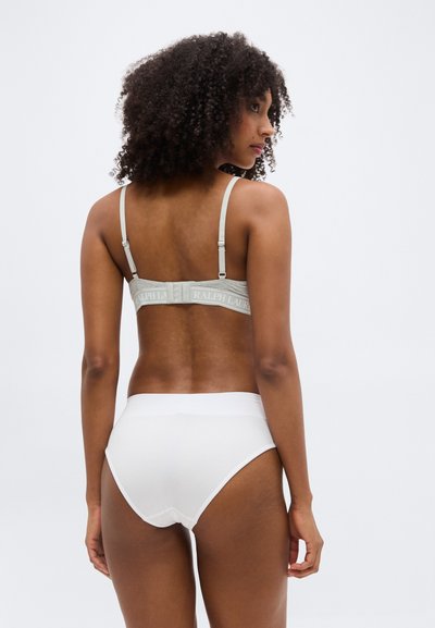 Femme aux cheveux bouclés portant un soutien-gorge gris et une culotte taille haute blanche, vue de dos sur un fond uni.