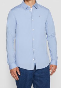 Uomo che indossa una camicia azzurro chiaro a maniche lunghe con un piccolo logo sul petto e jeans di denim blu scuro.