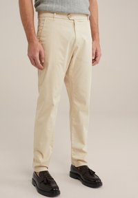 Pantaloni di cotone beige con taglio dritto, dotati di una chiusura frontale con bottone e tasche laterali. Abbinati a mocassini in pelle nera.