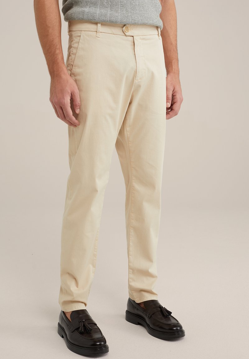 Pantaloni di cotone beige con taglio dritto, dotati di una chiusura frontale con bottone e tasche laterali. Abbinati a mocassini in pelle nera.