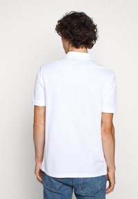Polo blanc en coton, doté d'un col classique et de manches courtes. Le dos présente une coupe droite avec des fentes latérales. Un jean bleu complète la tenue.
