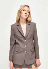 Brązowa marynarka w wzory z złotymi guzikami, z klapami i dwoma przednimi kieszeniami. Dopasowany krój z lekkim połyskiem i teksturowanym materiałem.