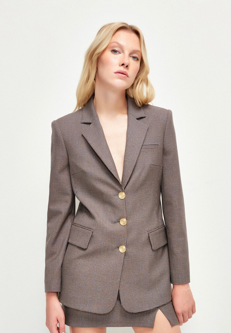 Brązowa marynarka w wzory z złotymi guzikami, z klapami i dwoma przednimi kieszeniami. Dopasowany krój z lekkim połyskiem i teksturowanym materiałem.