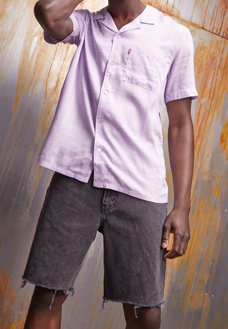 Man wearing a light purple short-sleeve button-up shirt and black frayed denim shorts against a textured rust-streaked wall.