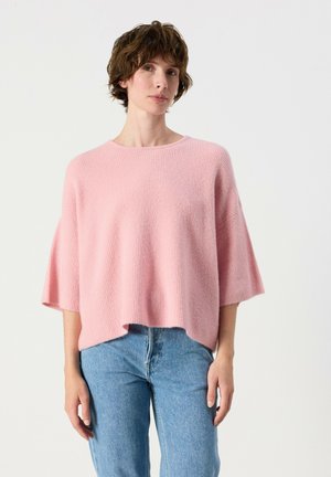Femme portant un pull rose ample à manches mi-longues et un jean bleu, debout devant un fond uni.