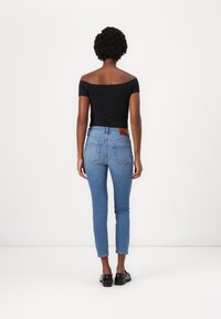 Zwarte off-the-shoulder top en blauwe slimfit jeans met een bruine leren patch op de achterkant van de taille. Model staat met de rug naar de camera tegen een witte achtergrond.