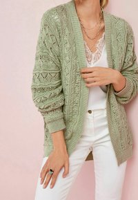 Hellgrüne Strickjacke mit filigranen Spitzenmustern, gerippten Bündchen und offenem Vorderteil, kombiniert mit weißem Tanktop und Hose.