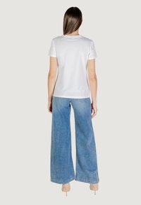 T-shirt bianca in cotone con maniche corte, abbinata a jeans a gamba larga di un blu chiaro, con una vestibilità casual e dettagli con orlo grezzo. Tacchi trasparenti visibili.