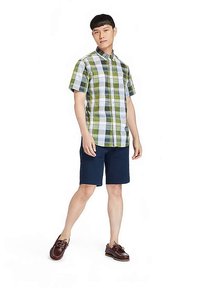 Kurzärmeliges kariertes Hemd in Grün, Weiß und Blau, kombiniert mit dunkelblauen Shorts und braunen Bootsschuhen, stehend vor einem schlichten Hintergrund.