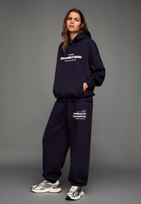 Sötét tengerészkék kapucnis pulóver és hozzáillő melegítőnadrág fehér feliratos részletekkel. A pulóveren elülső zseb található; a cipők fémes színűek, mintás kidolgozással.