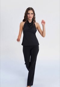 Frau mit welligem Haar, die ein ärmelloses schwarzes Oberteil und schwarze Hosen trägt, geht vorwärts vor einem schlichten hellen Hintergrund.