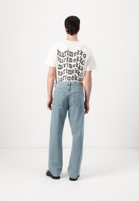 Tricou alb cu text negru suprapus care spune "marimekko" pe spate, asortat cu blugi largi de culoare albastru deschis și pantofi negri.