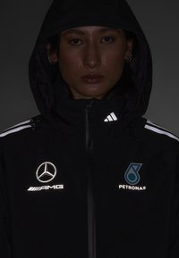 Sort hættejakke med reflekterende detaljer, som har logoer for Mercedes-Benz, AMG og Petronas. Indeholder lynlåse og et tekstureret stof.