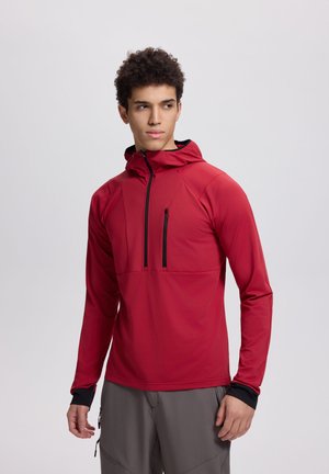 Jeune homme portant une veste à capuche rouge avec des fermetures éclair sur le devant et la poitrine, et un pantalon gris, regardant légèrement sur le côté sur un fond uni.