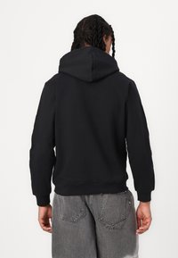 Svart hoodie med huva, ribbade muddar och fåll. Tyget verkar mjukt och slätt. Bärs med grå, lätt slitna shorts.