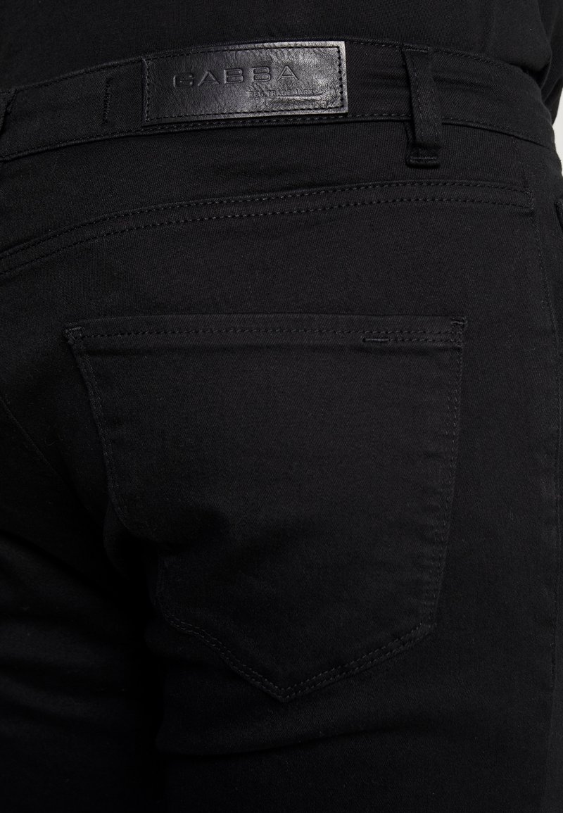 Vaqueros de mezclilla negra con un bolsillo trasero visible, etiqueta de cuero con "GABBA" y un acabado de tela suave y texturizado.
