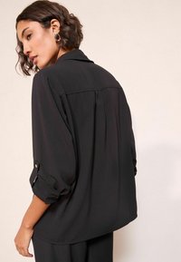 Blusa nera con una vestibilità rilassata, maniche lunghe arrotolate, con un sottile plissettatura sul retro e dettagli con bottoni sui polsini. Texture morbida.