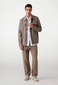 Giacca marrone chiaro con bottoni, con due tasche sul petto, indossata sopra una camicia bianca, abbinata a pantaloni coordinati e scarpe da ginnastica chiare. Tessuto dalla texture liscia.