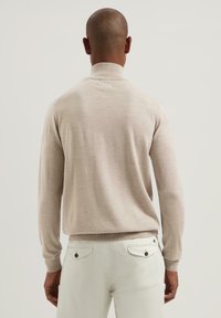 Beige rolák s dlouhými rukávy, měkkou texturou a žebrovanými manžetami, kombinovaný se světle barevnými kalhotami, zobrazený z hladkého zadního pohledu.