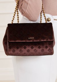 Brązowa aksamitna torebka z tłoczonymi wzorami, złotym łańcuszkiem na ramię i złotym logo marki. Zamykana klapką o wyraźnej, strukturalnej formie.