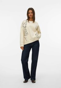 Felpa crema con design in arte ligneo nero, vestibilità comoda, polsini a coste, abbinata a jeans a gamba larga blu scuro e scarpe marroni.