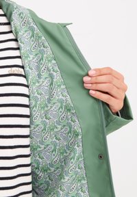 Grön regnjacka med paisleymönstrad foder i nyanser av grönt och grått. Har dragkedjestängning och en hand som håller jackan öppen.