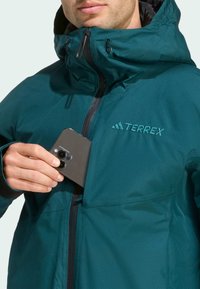 Teal weatherproof jacket with a zippered chest pocket, featuring embroidered "TERREX" logo and black accents on cuffs and zipper.