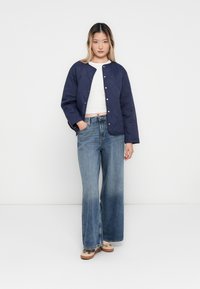 Junge Frau, die eine marineblaue Steppjacke über einem weißen Oberteil, weite blaue Jeans und beige Sneakers trägt, vor einem einfarbigen Hintergrund steht.
