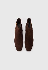 Botas de tobillo de ante marrón con un diseño de punta cuadrada, que cuentan con un cierre de cremallera lateral y acentos de hardware dorado. Textura suave en general.