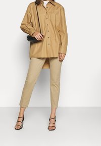 Luźna, jasno-brązowa koszula na guziki z kieszenią z przodu, zestawiona ze zwężanymi beżowymi spodniami i czarnymi sandałami na paski. Akcesorium w postaci czarnej torebki na ramię.