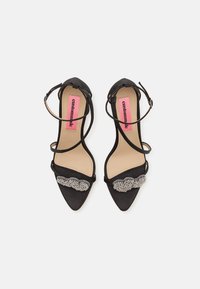 Black suede high-heeled sandals with pointed toes, strappy design, and a metallic decorative detail across the front. Pink insole branding.