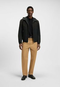 Chaqueta negra con cremallera y capucha, combinada con una camiseta polo oscura y pantalones beige. Calzado: mocasines negros. Ajuste limpio y moderno.