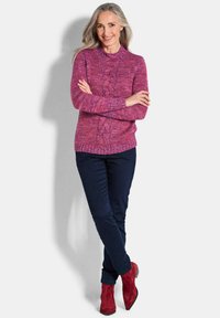 Strickpullover mit rundem Ausschnitt in bunten Pinktönen, mit einem Zopfdesign. Kombiniert mit dunklen Jeans und roten Wildleder-Stiefeletten.