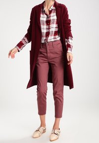 Long burgundy coat over a red-and-white plaid button-up shirt, paired with slim maroon trousers and cream shoes with straps.