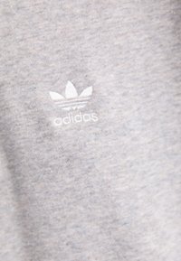 Tejido de algodón gris con un pequeño logo blanco de Adidas que presenta tres rayas en forma de hoja por encima del nombre de la marca. Textura suave con un sutil patrón jaspeado.